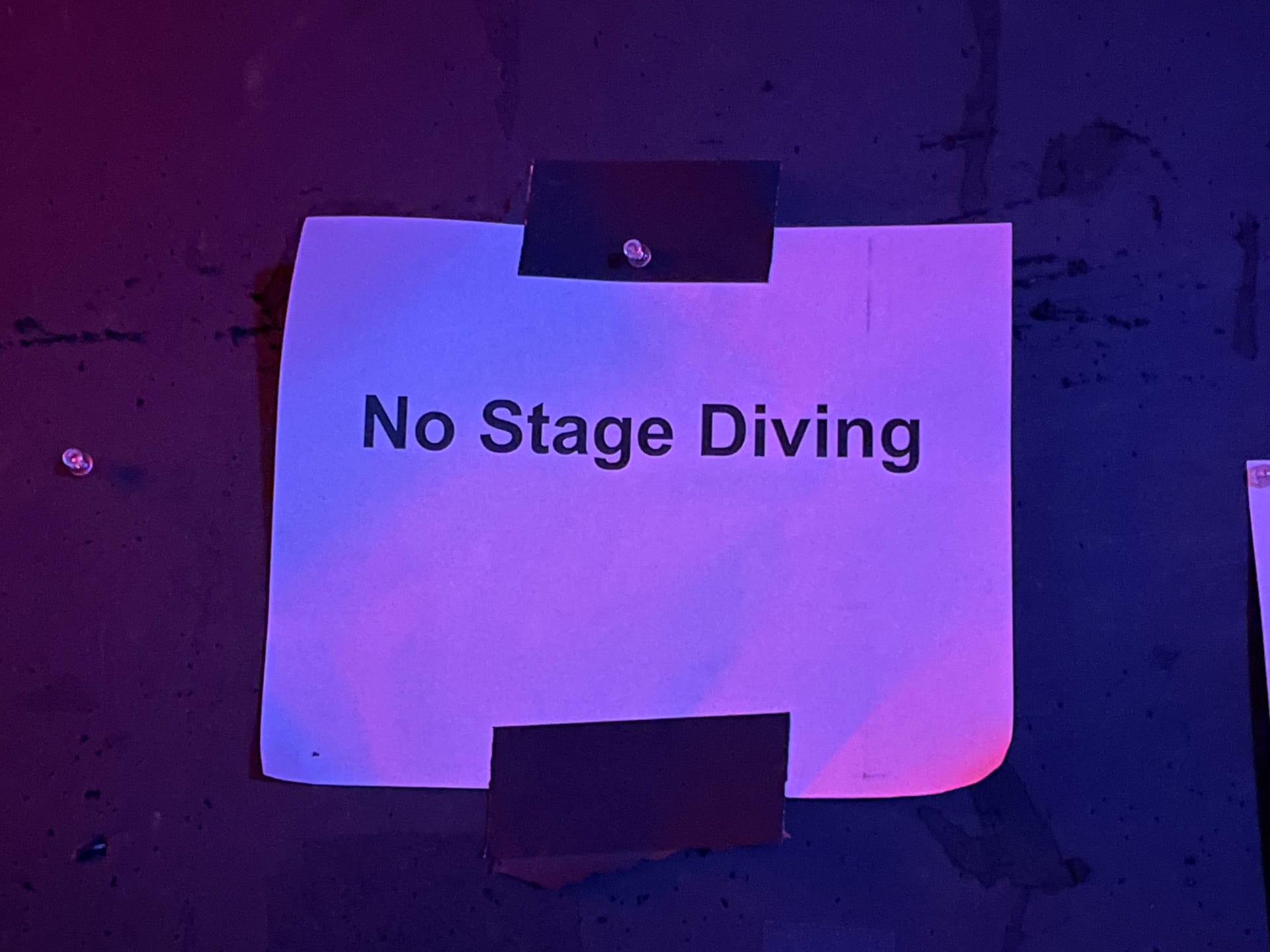A white piece of paper with the words "No Stage Diving" printed on it. It is attached to the wall with black gaffers tape.. 